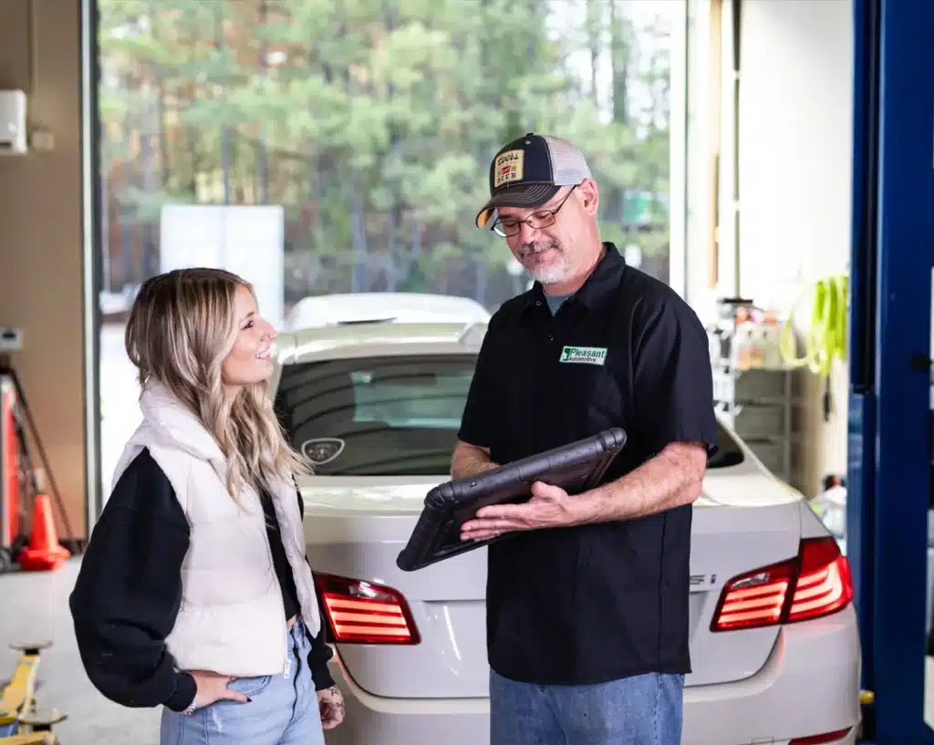 A woman and a man with a digital tablet standing in a car repair shop by the rear of a white car.