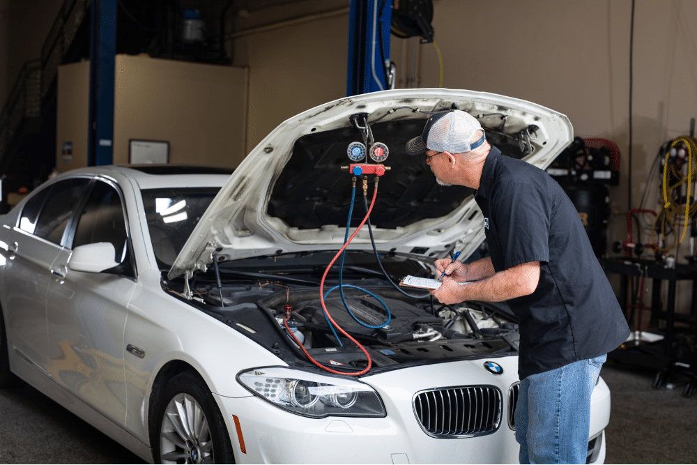 a/c problems, Auto Repair Services in Wake Forest, NC at Pleasant Automotive. Mechanic inspecting a BMW with the hood open, using A/C diagnostic gauges and writing notes on a clipboard in a service bay.